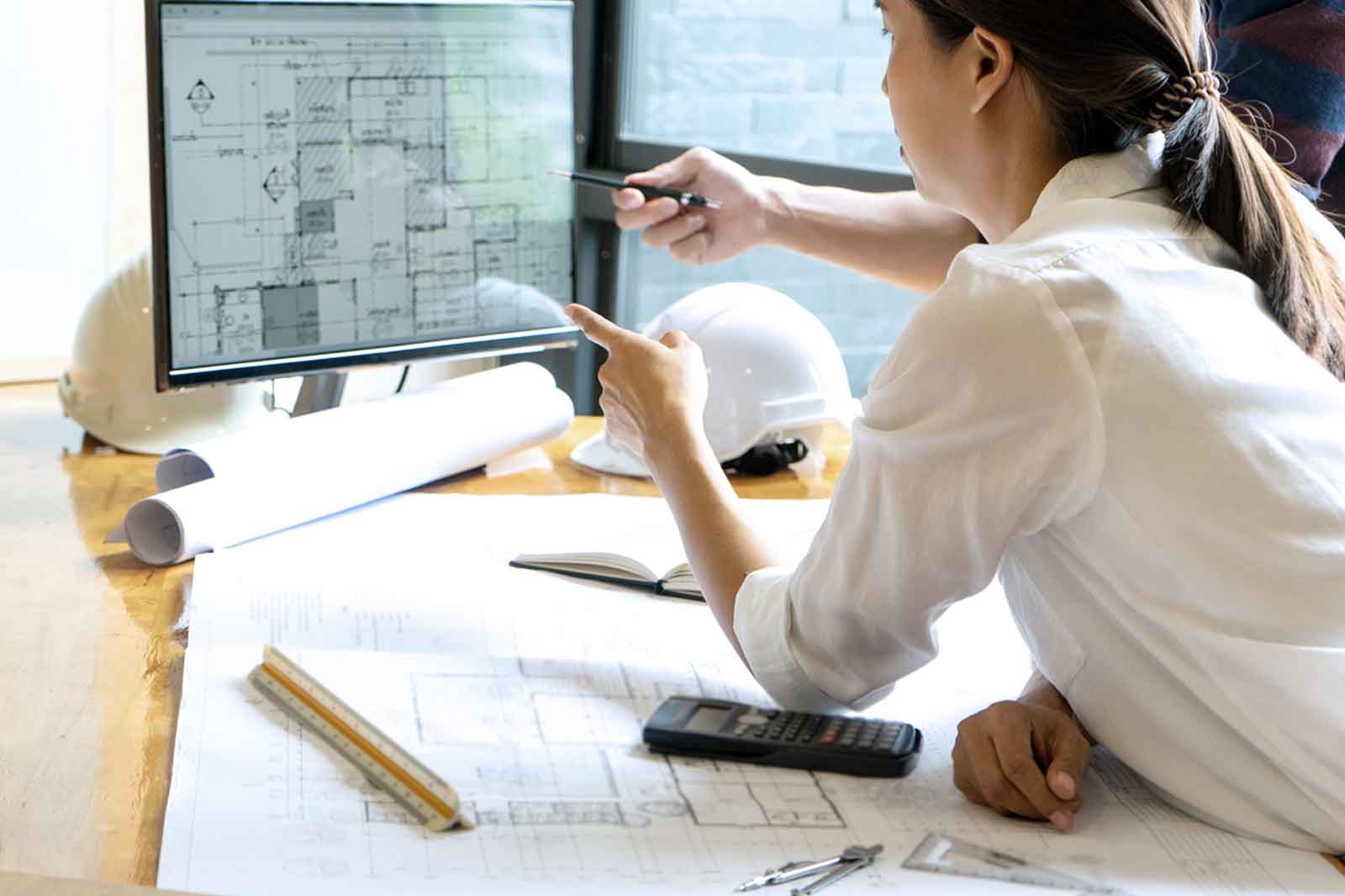 Eine Bauzeichnerin vergleicht ihren technischen Bauplan mit einer handgezeichneten Skizze an ihrem Arbeitsplatz.